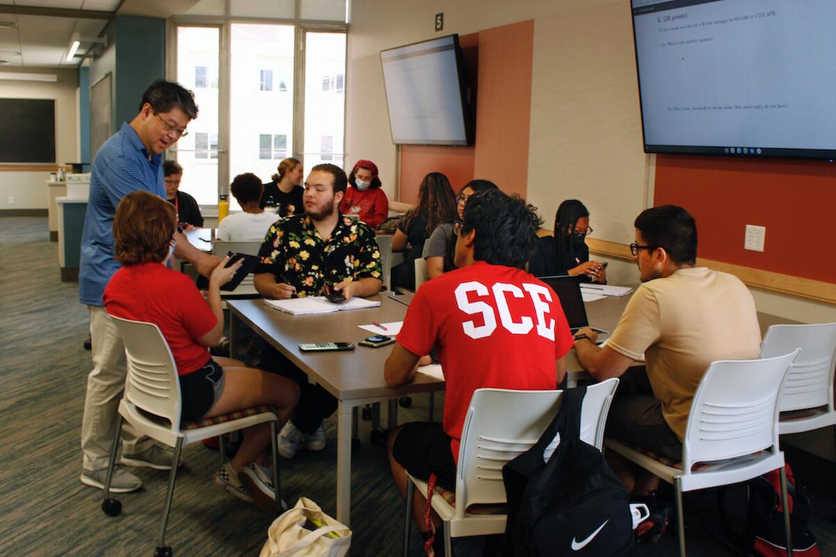 A standing instructor speaks to young adult students seated around a table