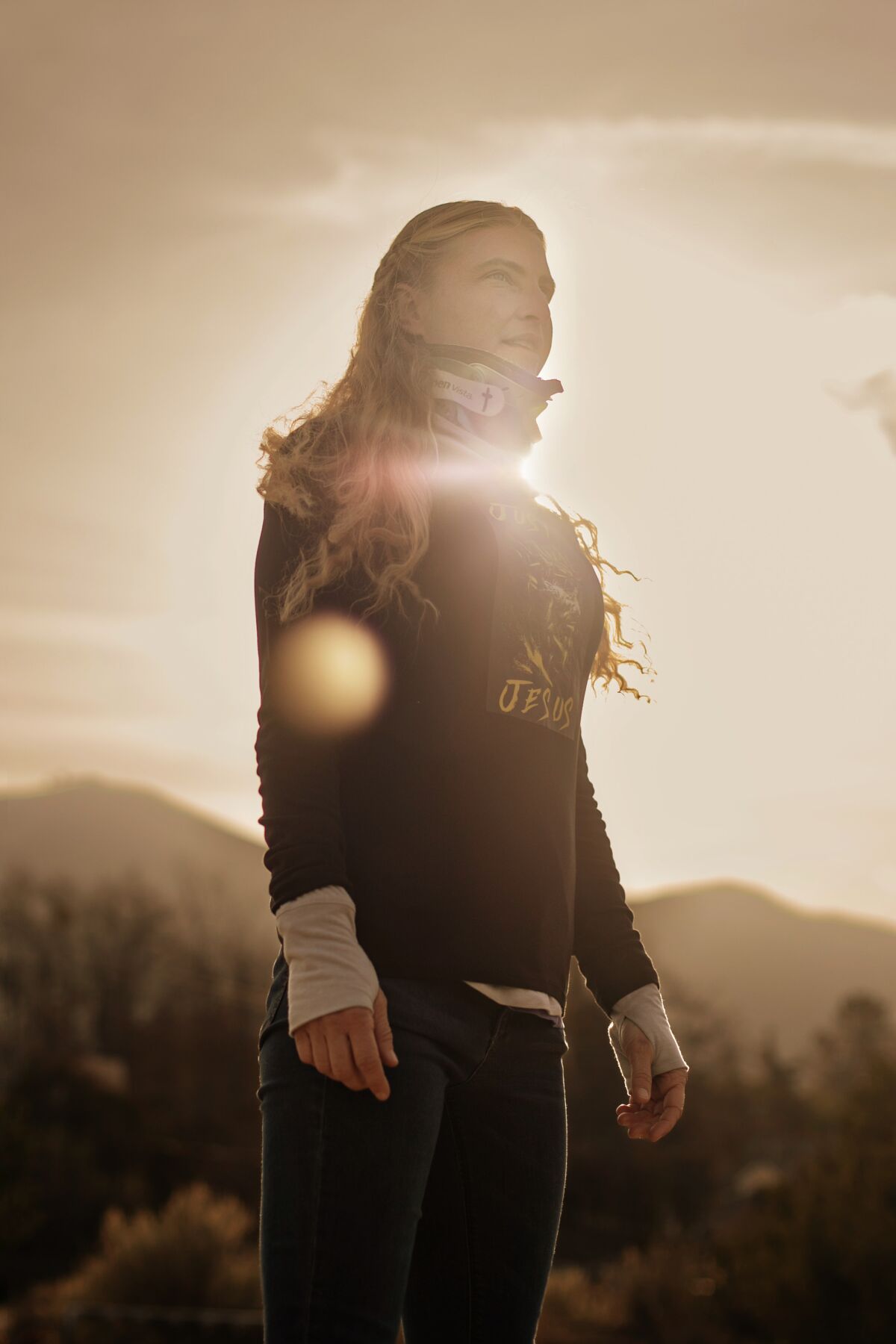 A vertical view of a blonde woman in a 3/4 view looking resolute, backlit by a setting sun.