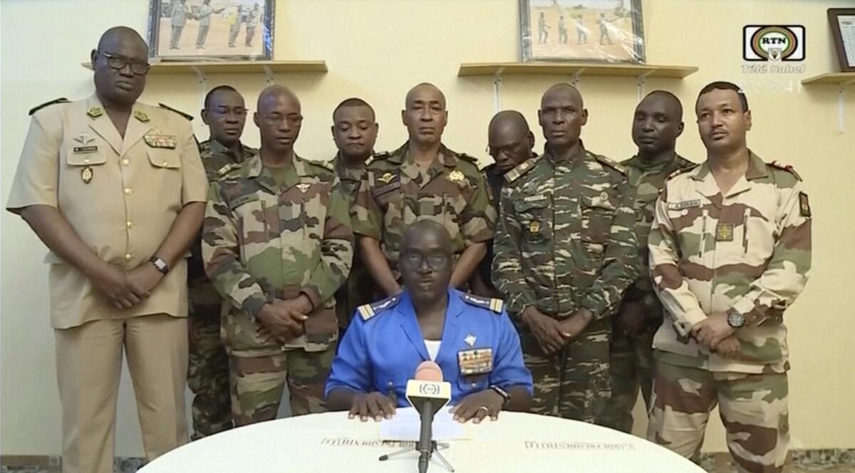 Col. Maj. Amadou Abdramane sits at a table behind a microphone while military officers stand behind him