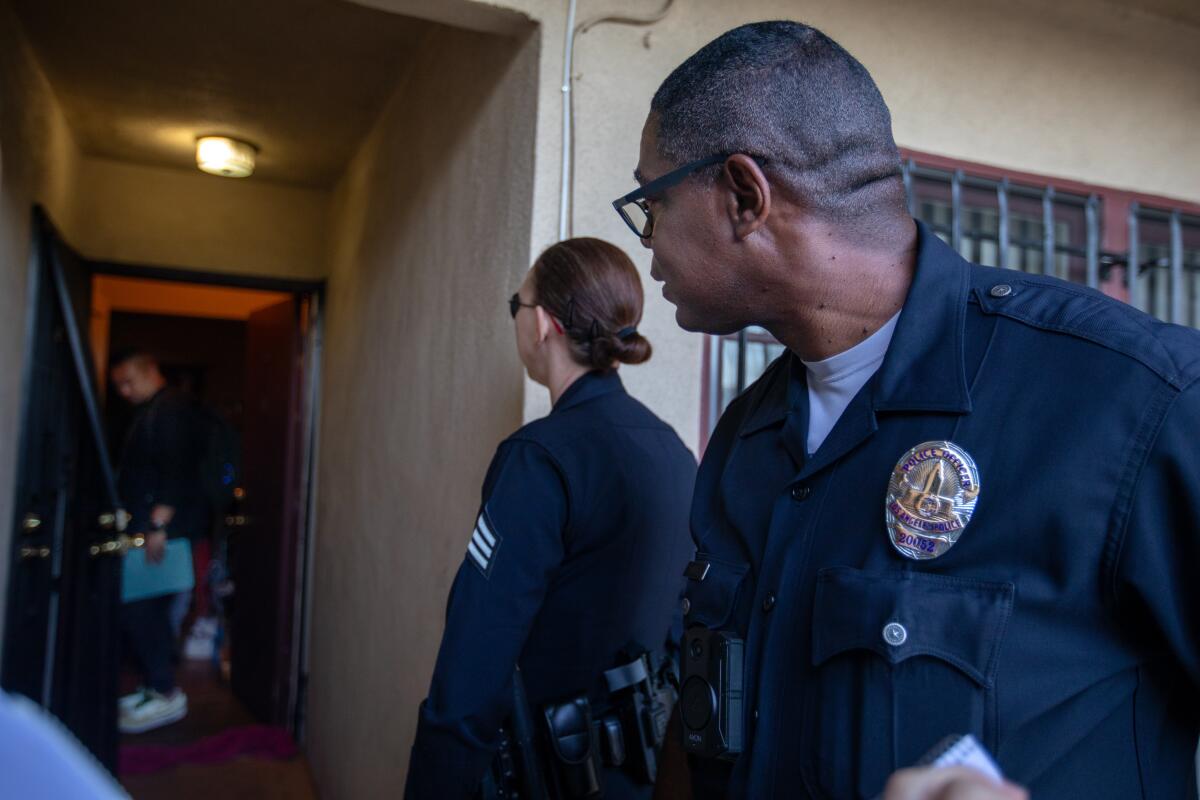 Two police officers look down a corridor at a man standing at an apartment door