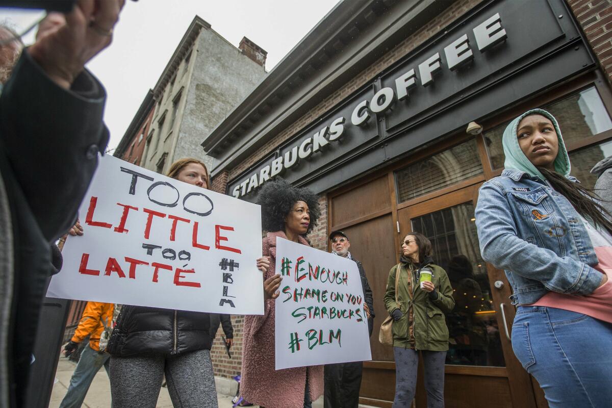 Several people protest outside a Starbucks store, some with Black Lives Matter signs, one reading "Too little/Too latte."