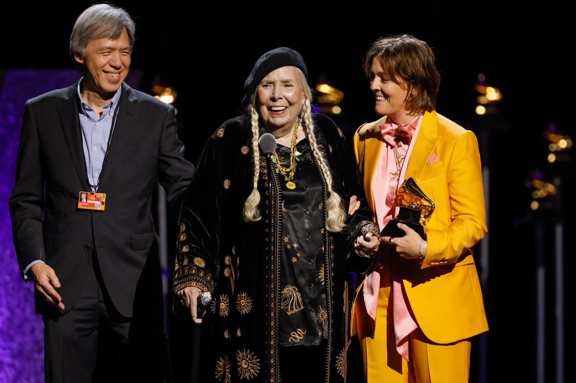 A man stands to the left of Joni Mitchell and Brandi Carlile to her right as Mitchell accepts a Grammy