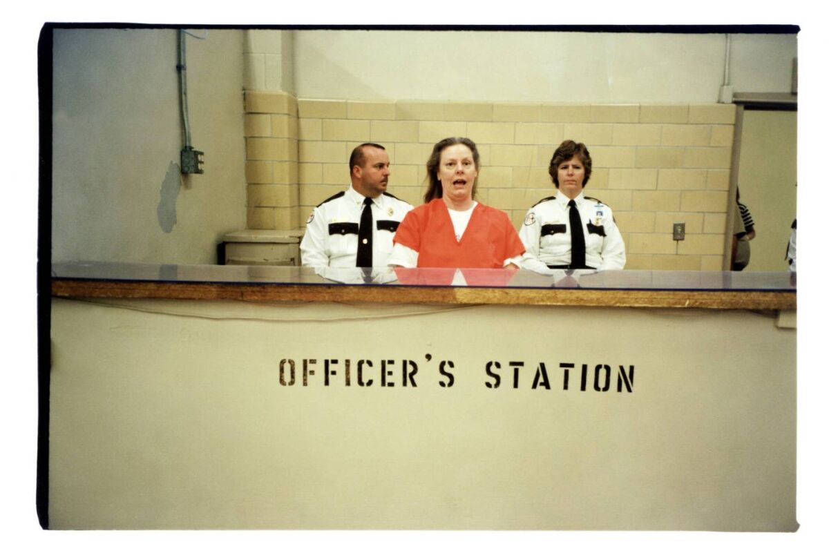 Aileen Wuornos in prisoner orange garb stands with two uniformed officers behind a counter marked “Officer’s station.” 