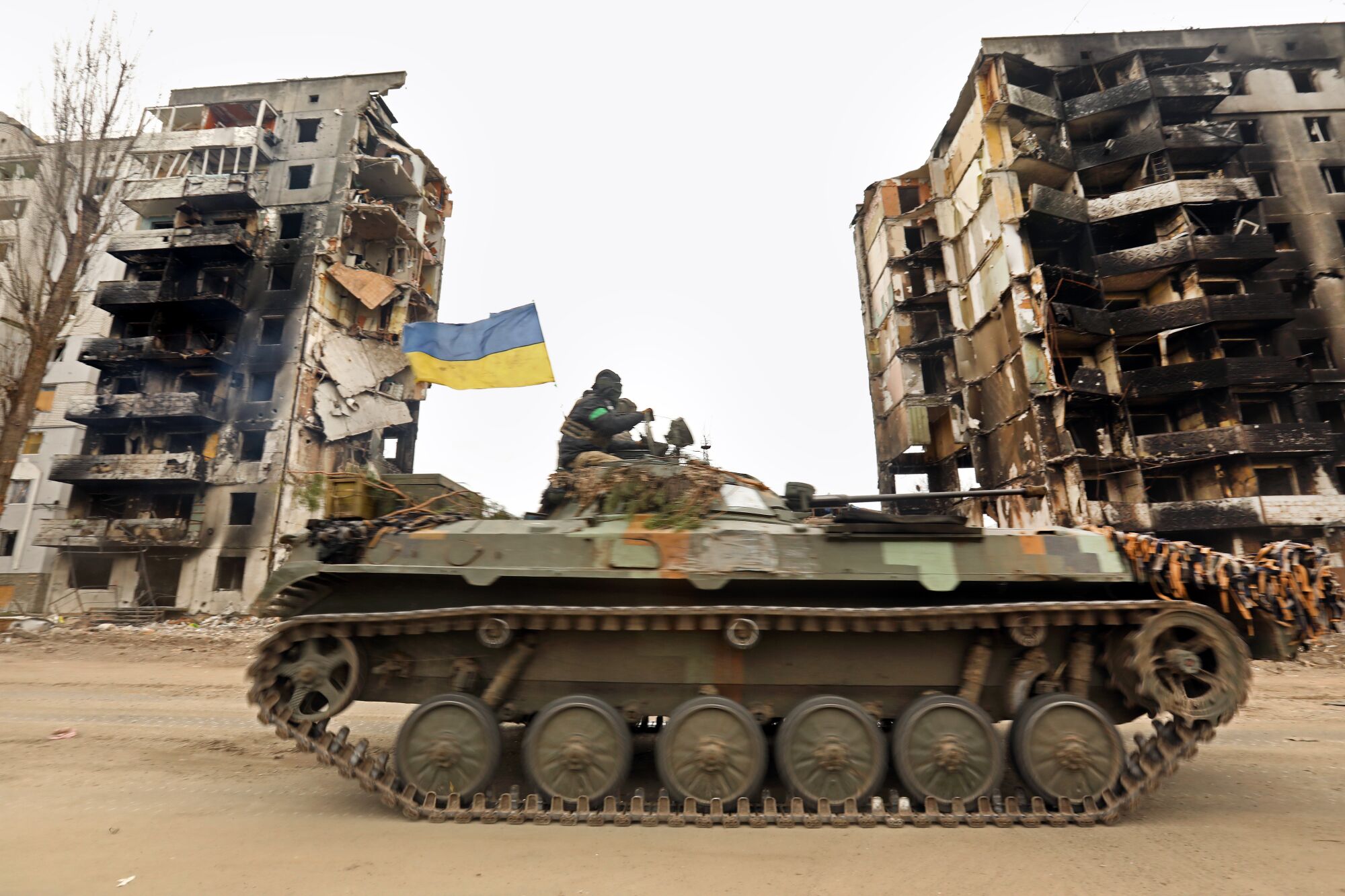 A tank with the blue-and-yellow Ukrainian flag rolls past burned-out buildings.