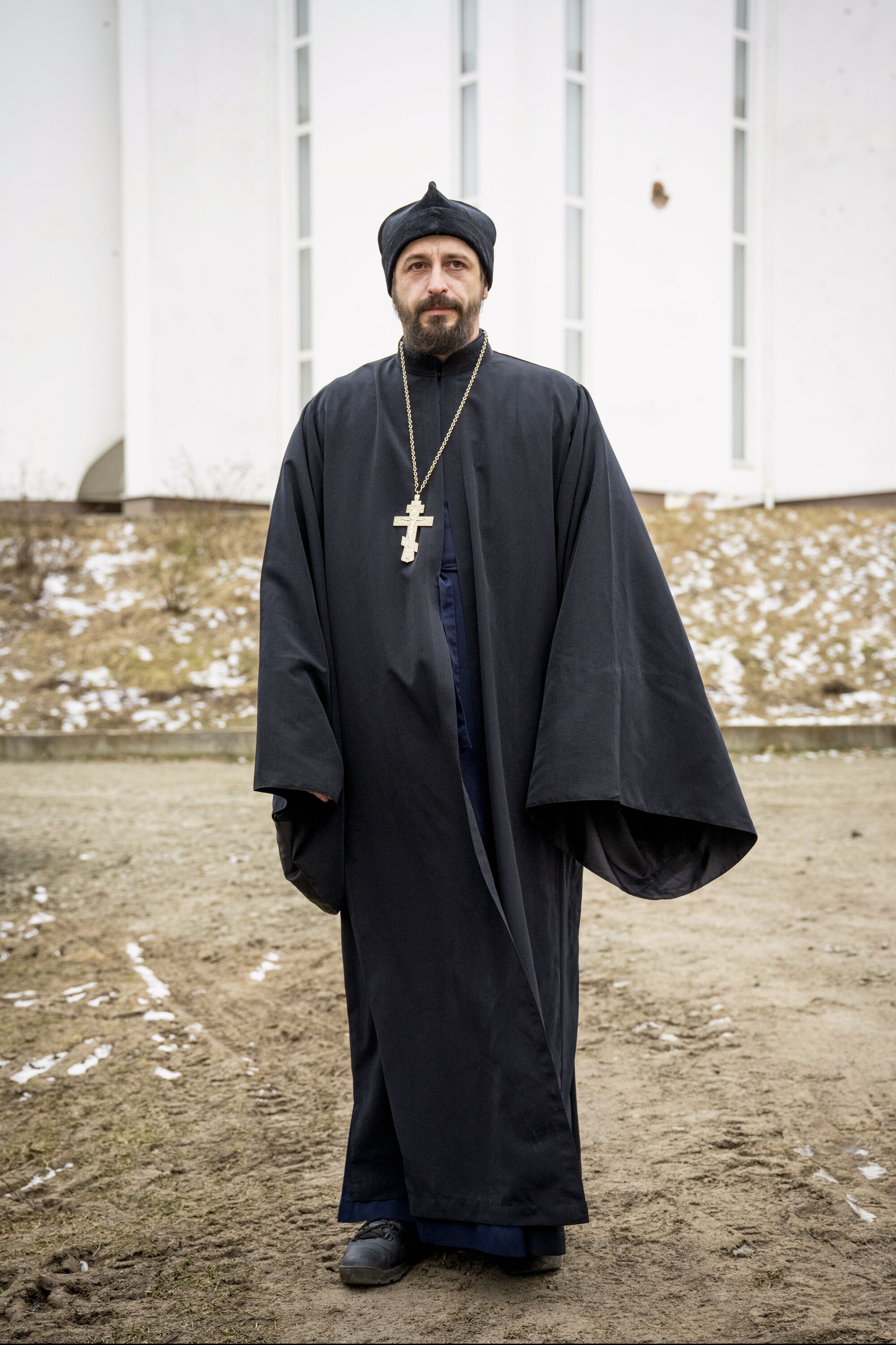 एक पुजारी, काले वस्त्र, काली टोपी पहने, और एक श्रृंखला पर एक बड़ा क्रॉस पहने हुए, एक चित्र के लिए प्रस्तुत करता है।