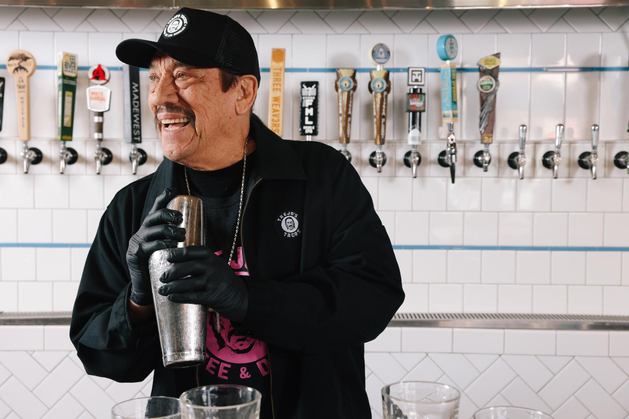 Danny Trejo smiles and holds a cocktail shaker in front of a row of beer taps