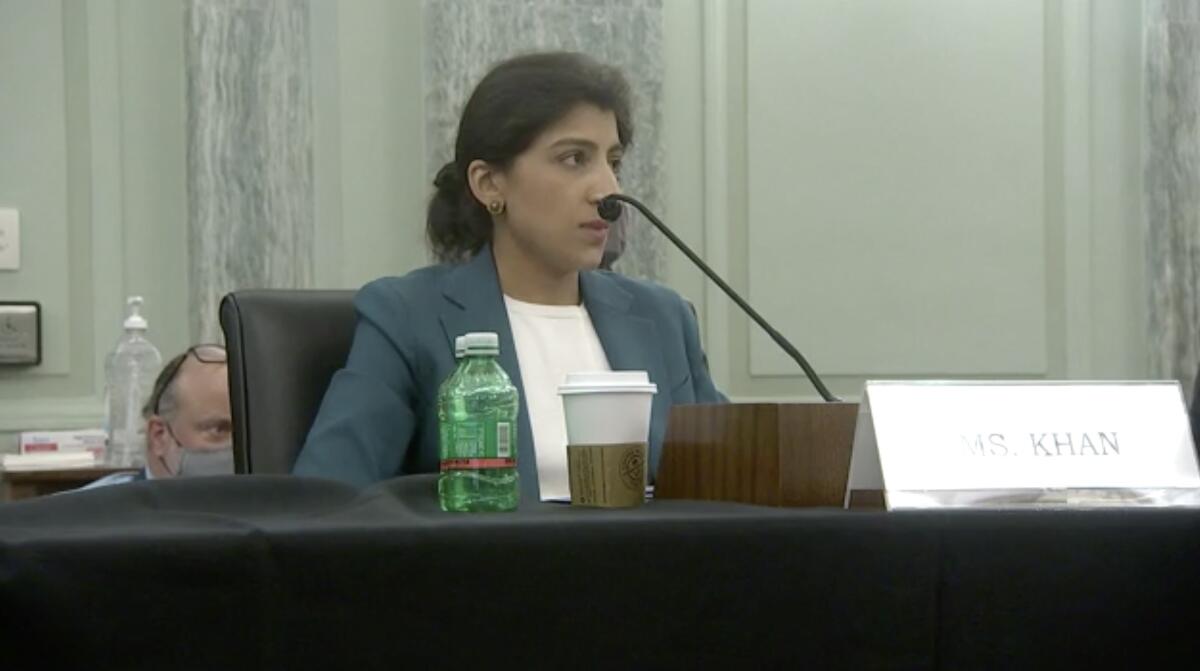 Lina Khan sits at a table, speaking into a microphone.