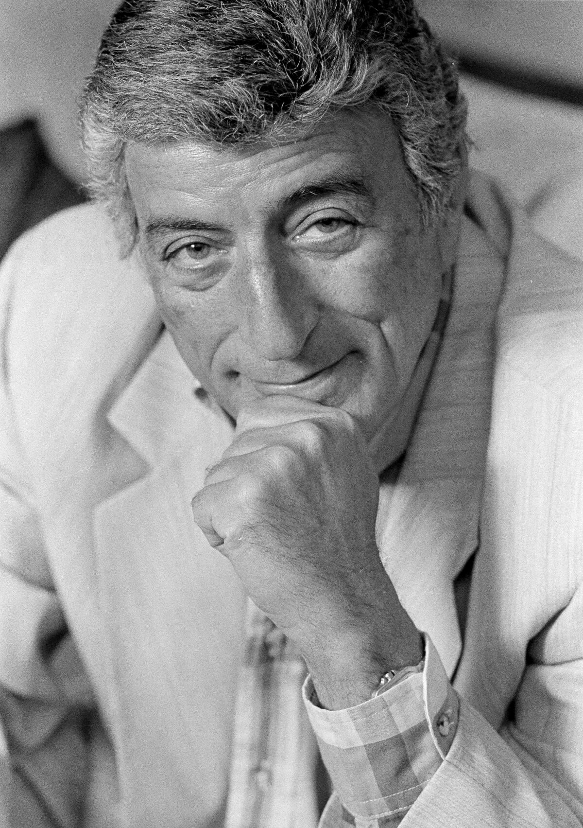 A vertical black-and-white frame of Tony Bennett looking at the camera while resting his chin on his left fist, smiling