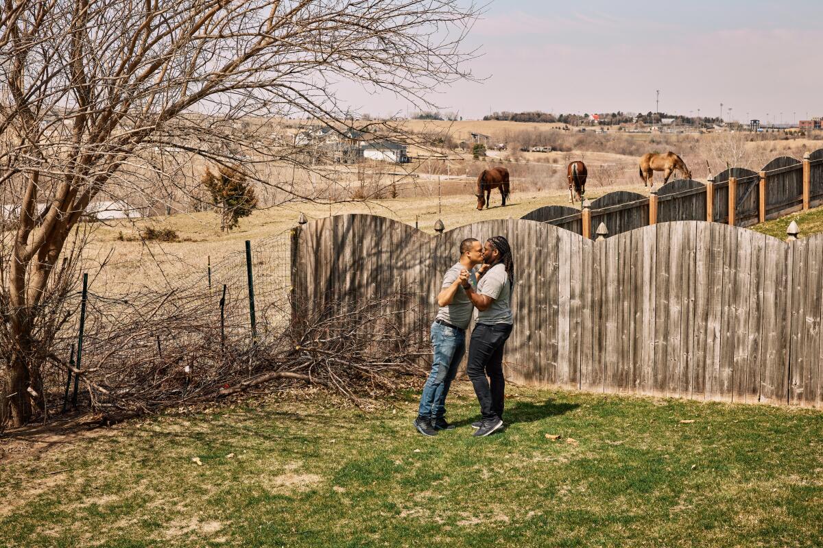 Two men kiss in a backyard with horses over the fence.
