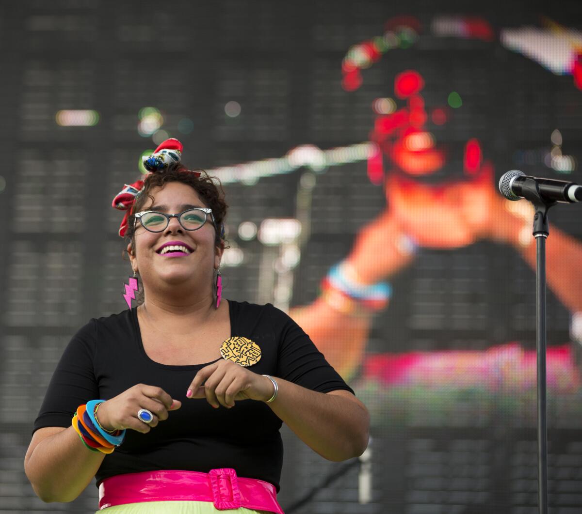 A smiling woman in glasses wears colorful dangling earrings, bangles and a pink belt.