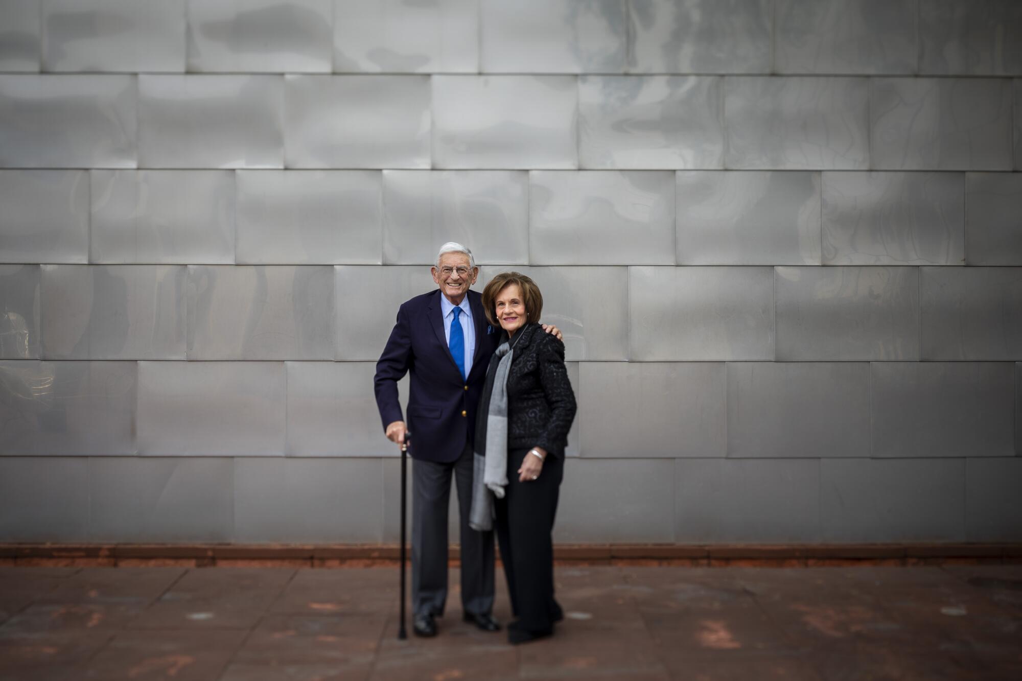 Eli Broad, leaning on a cane, puts an arm around wife Edythe as they stand in front of a wall of gray metal panels.