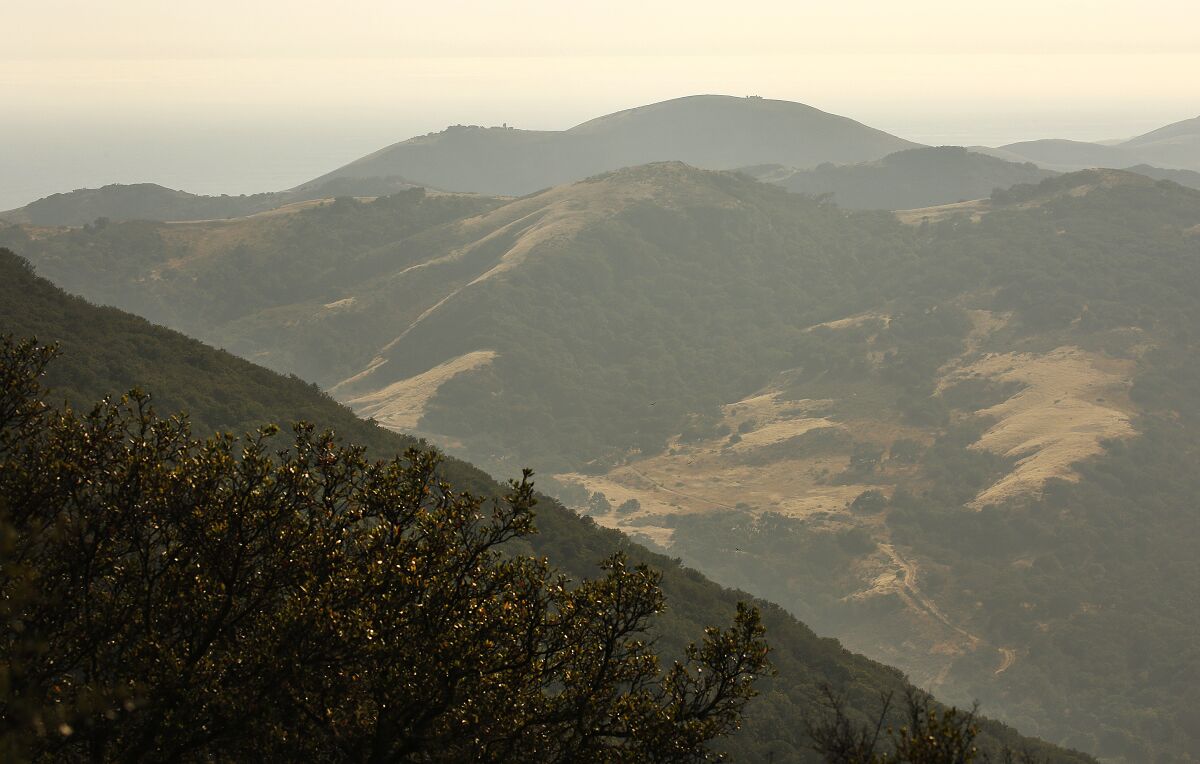 Green and gold hills seen from a mountaintop stretch toward a hazy horizon