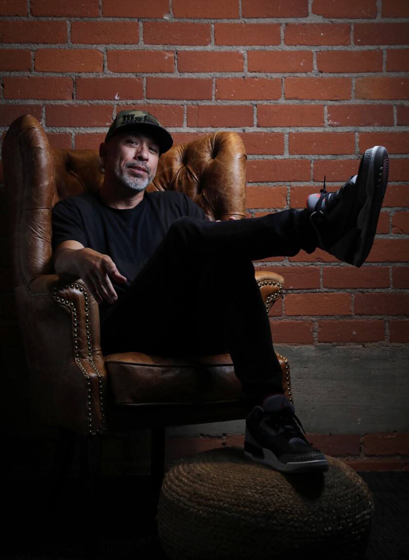 A man sits cross-legged in a leather chair in front of a brick wall.