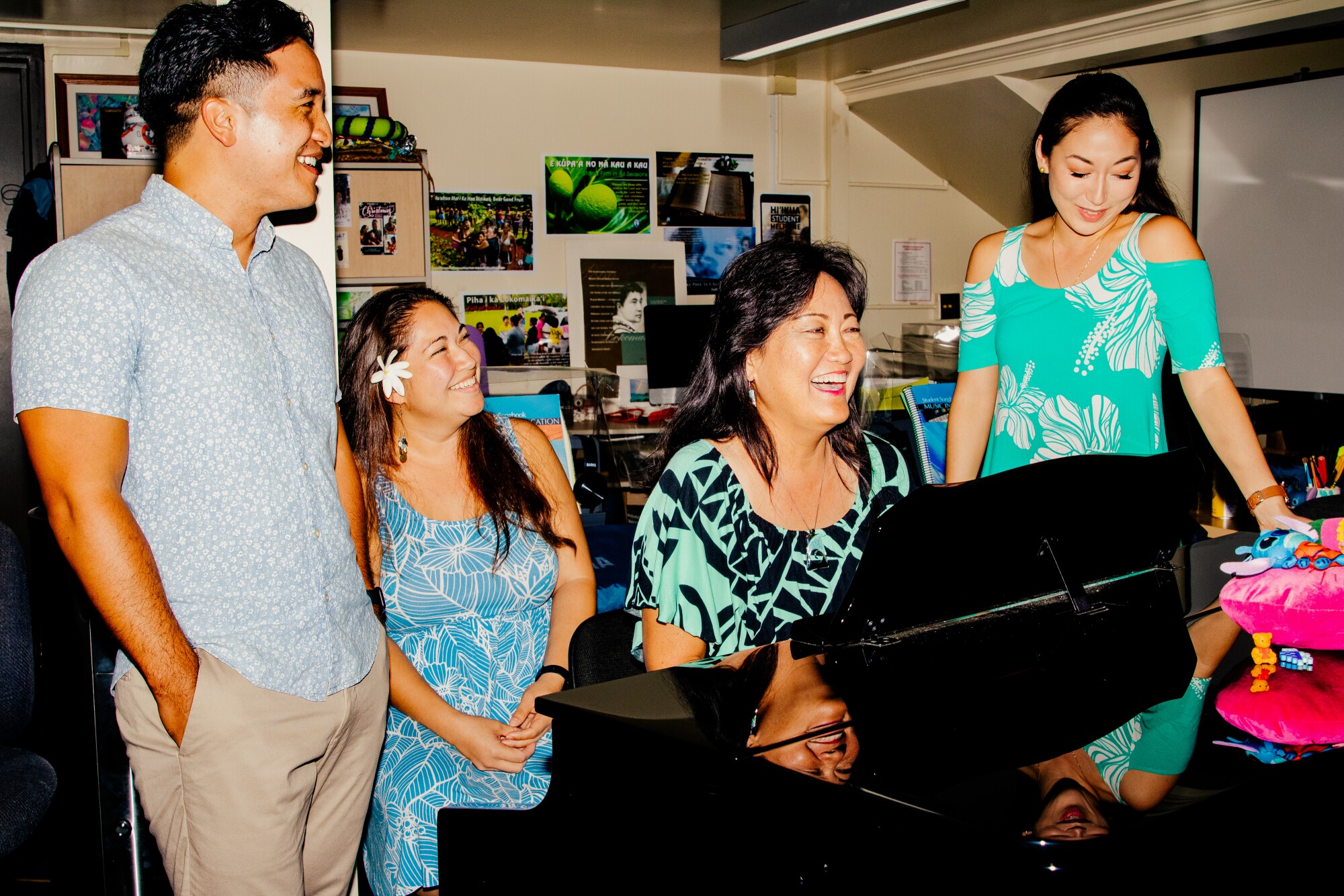 A man and four women smiling and gathered around a piano in a classroom