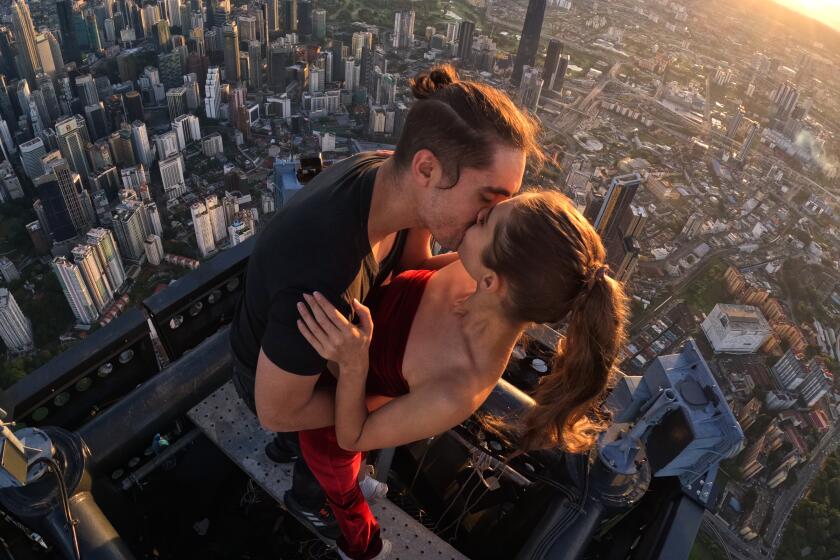 A couple embraces while perched on the rooftop of a building with thee ground far below.