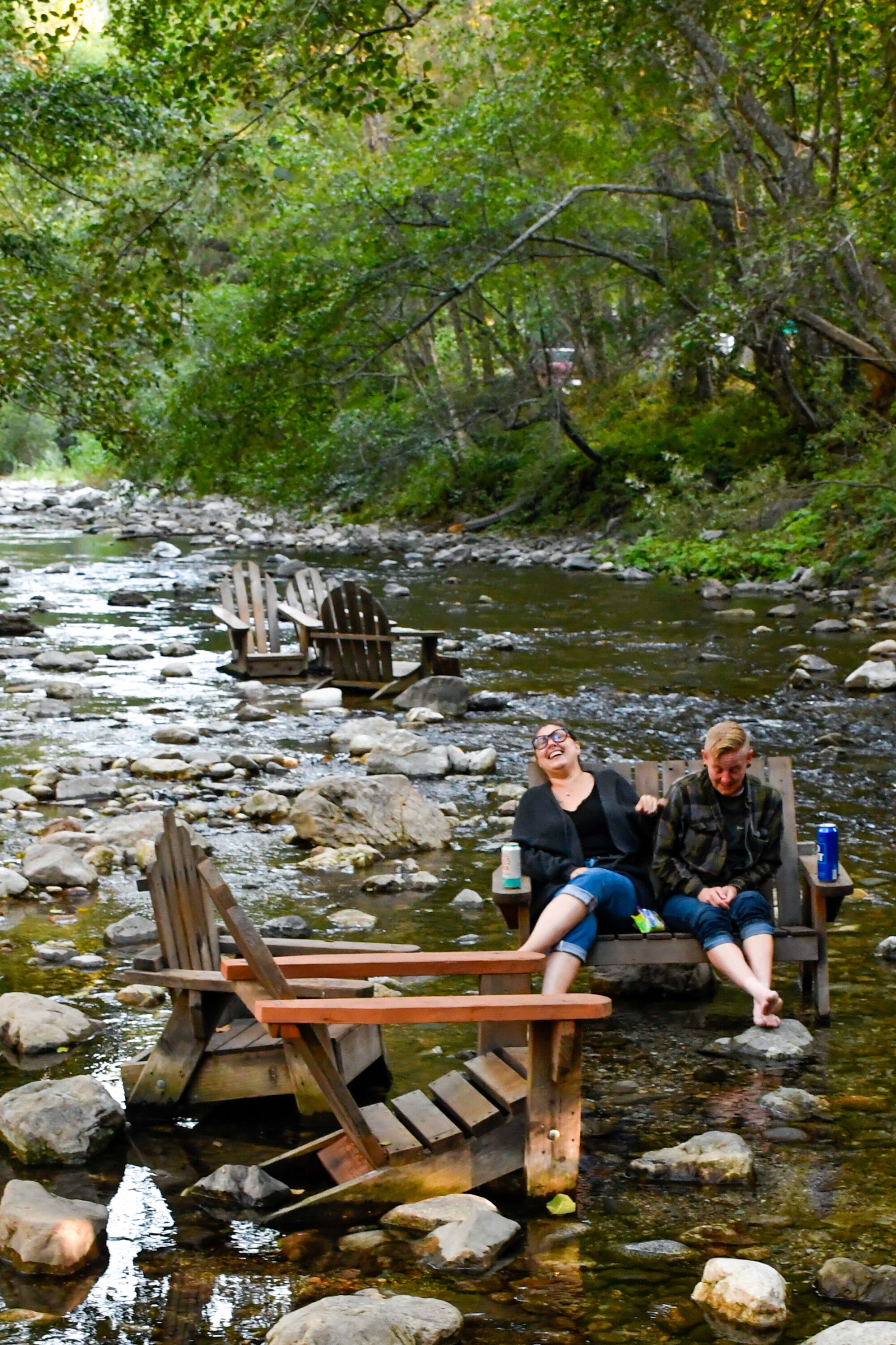 Two people sit in wooden chairs in the middle of a shallow part of a forested river.
