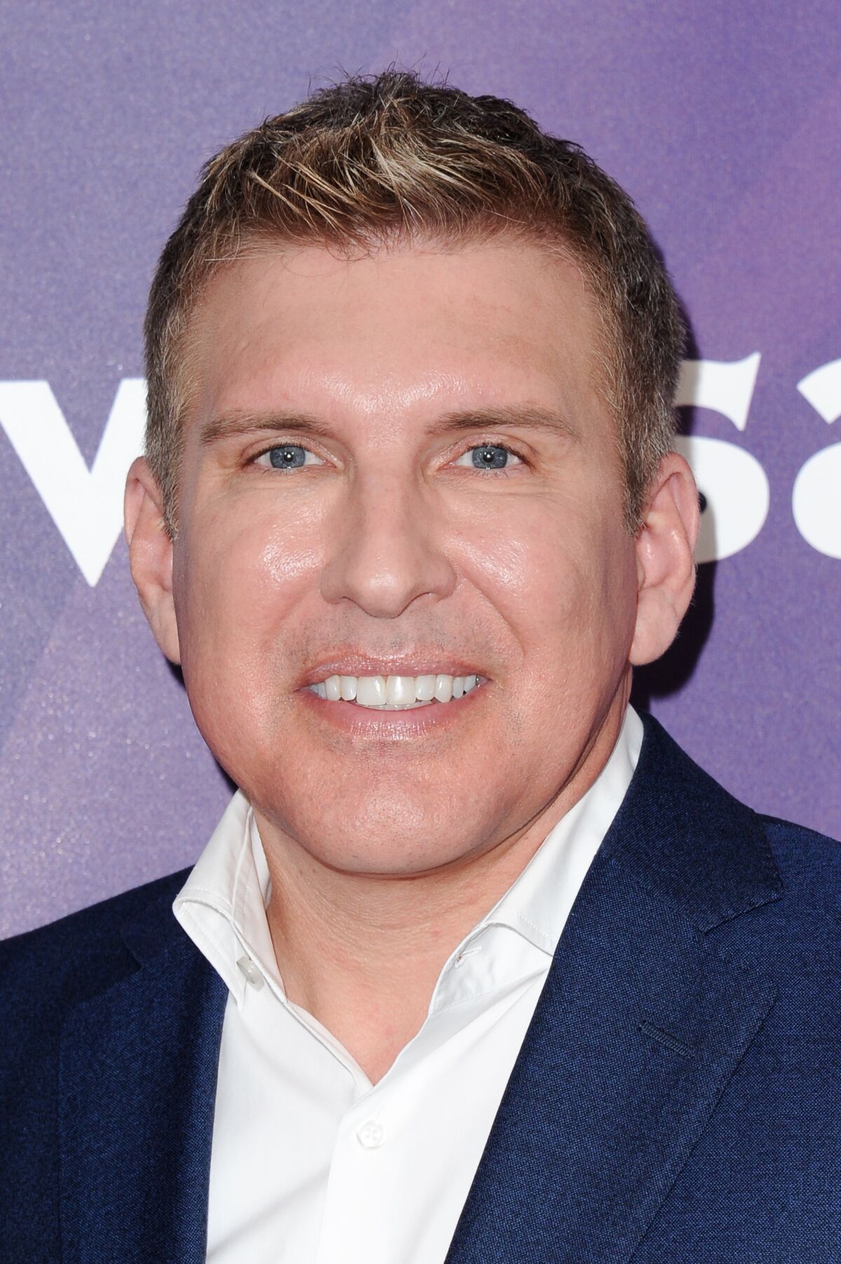 A man with short blond hair in a white button down shirt and dark blue blazer smiling