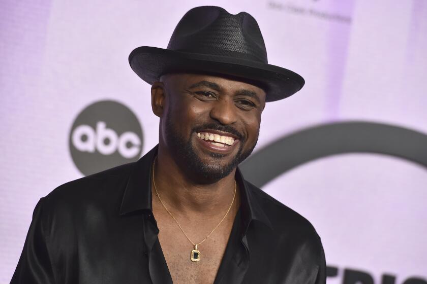 Wayne Brady smiles while wearing a black fedora and a black button up that reveals his chest and a gold necklace