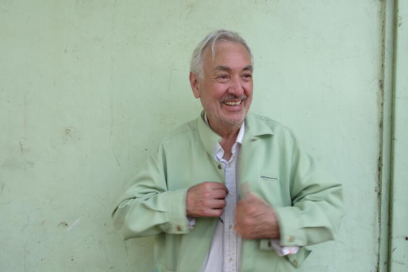 A man with grey hair wearing a mint green jacket smiles before a mint green wall