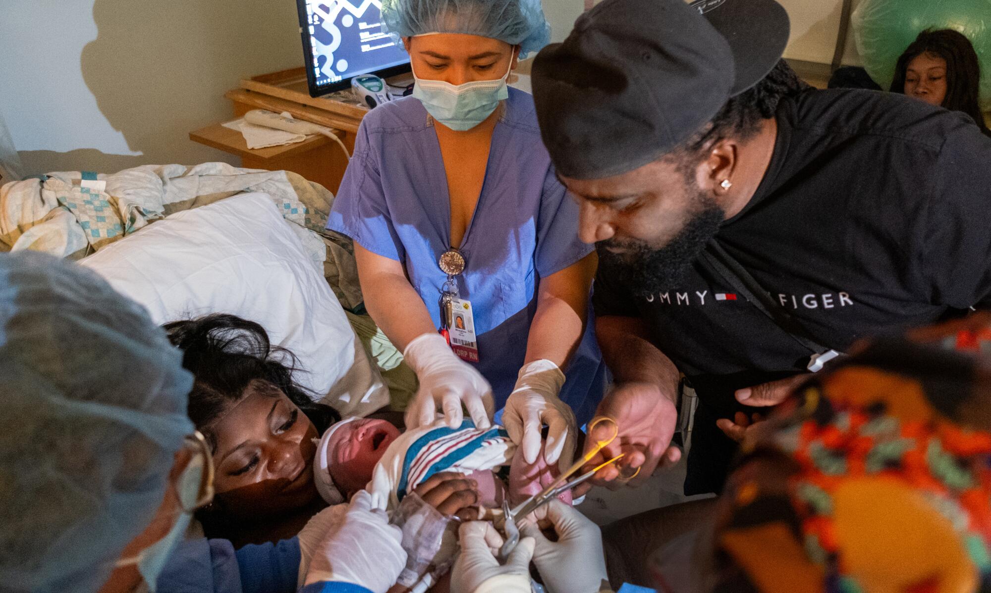 A man cuts the umbilical cord of his child as the mother lies in her hospital bed flanked by nurses.