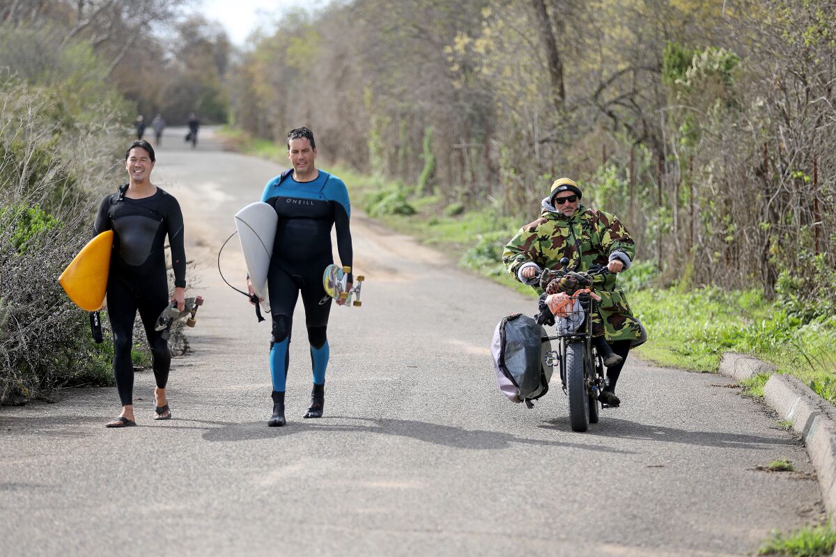 A man on an e-bike passes two surfers walking on a trail.