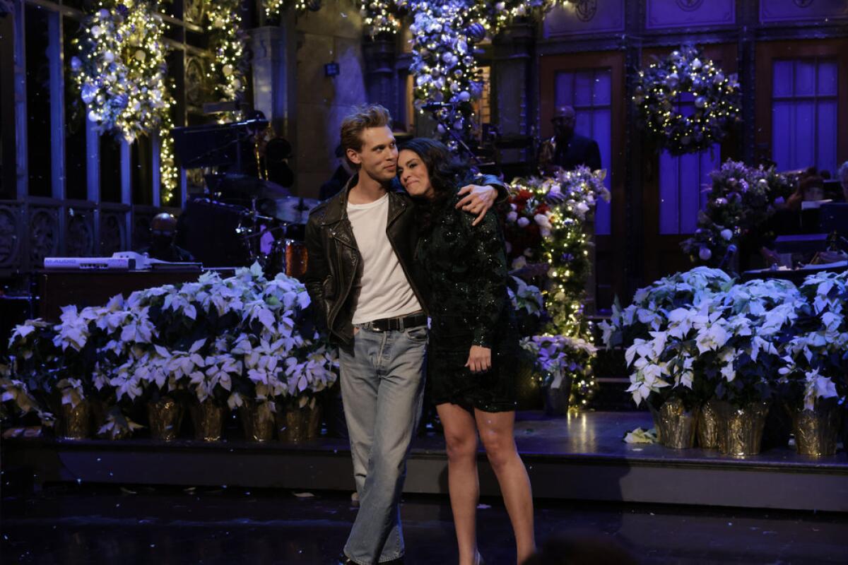 A blond man in a black leather jacket hugs a woman on a flower-covered stage