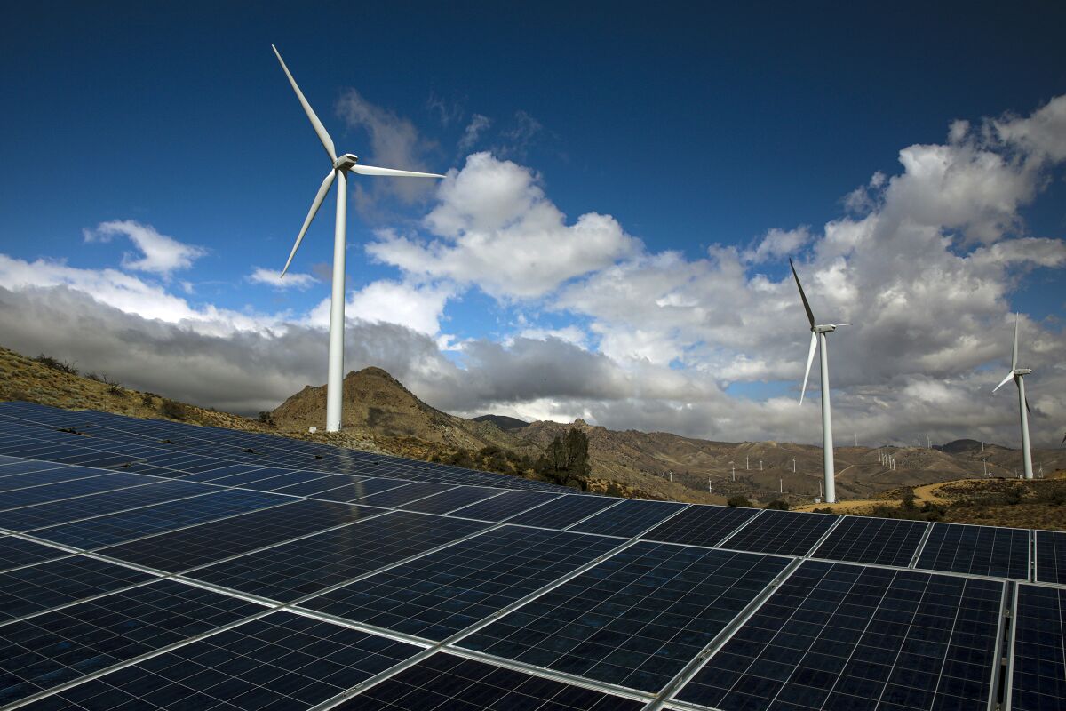 Solar panels are seen in the foreground, with wind turbines, mountains and blue sky and clouds in the background.