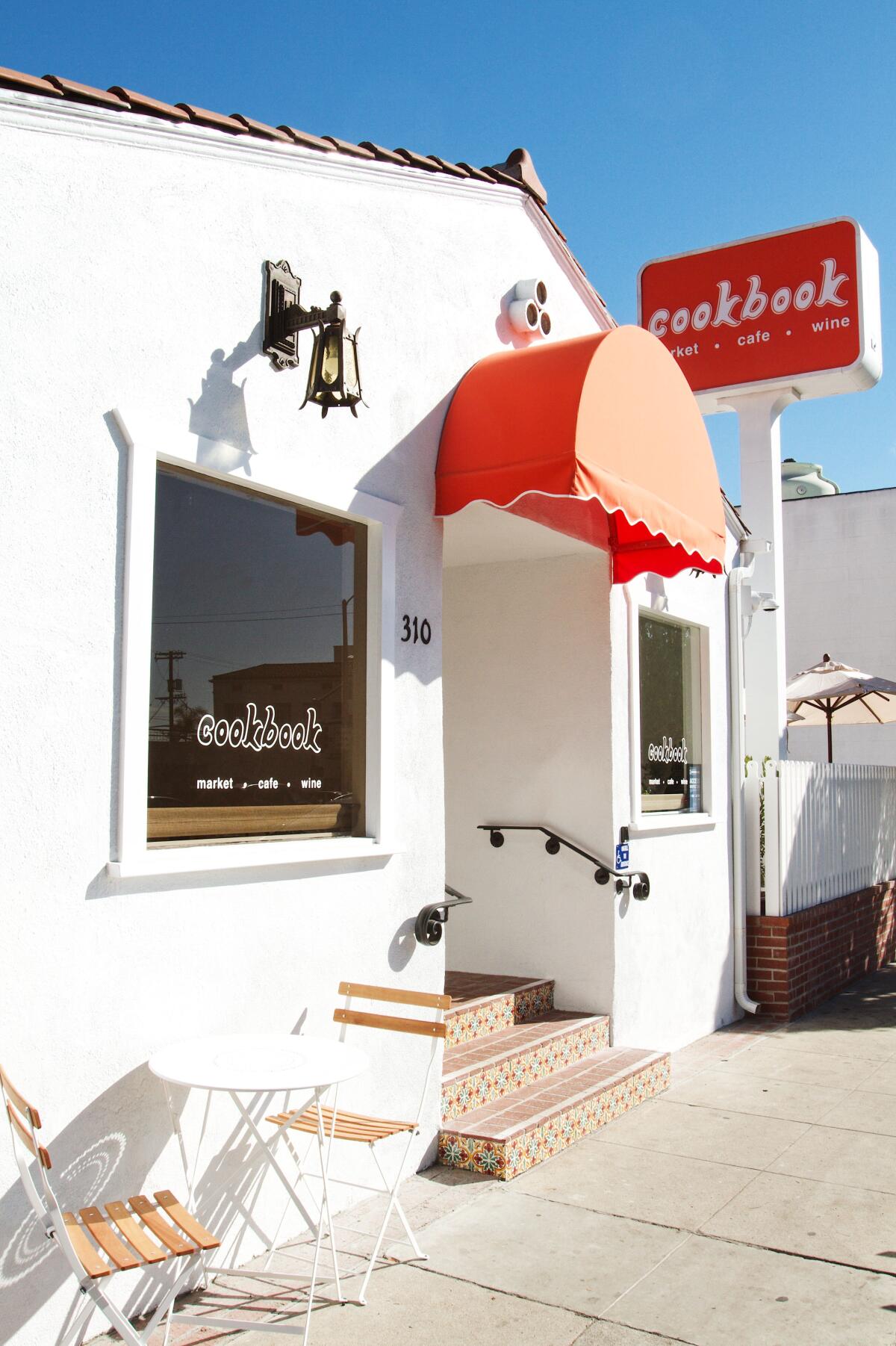 An exterior of the all-white Cookbook Market in Larchmont with orange signage and awning.