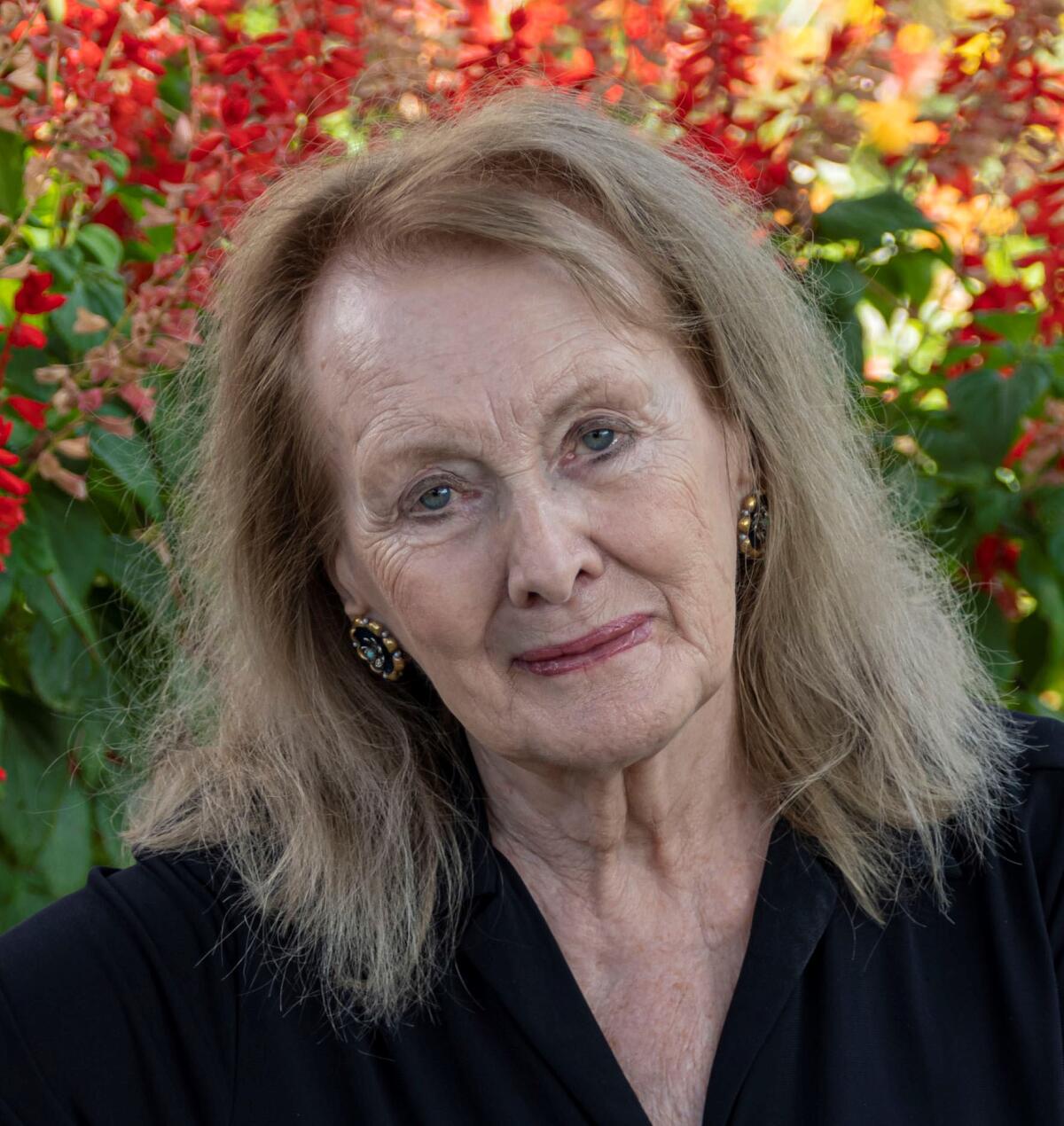 An older woman with light blond hair and an open black shirt with red flowers in the background.