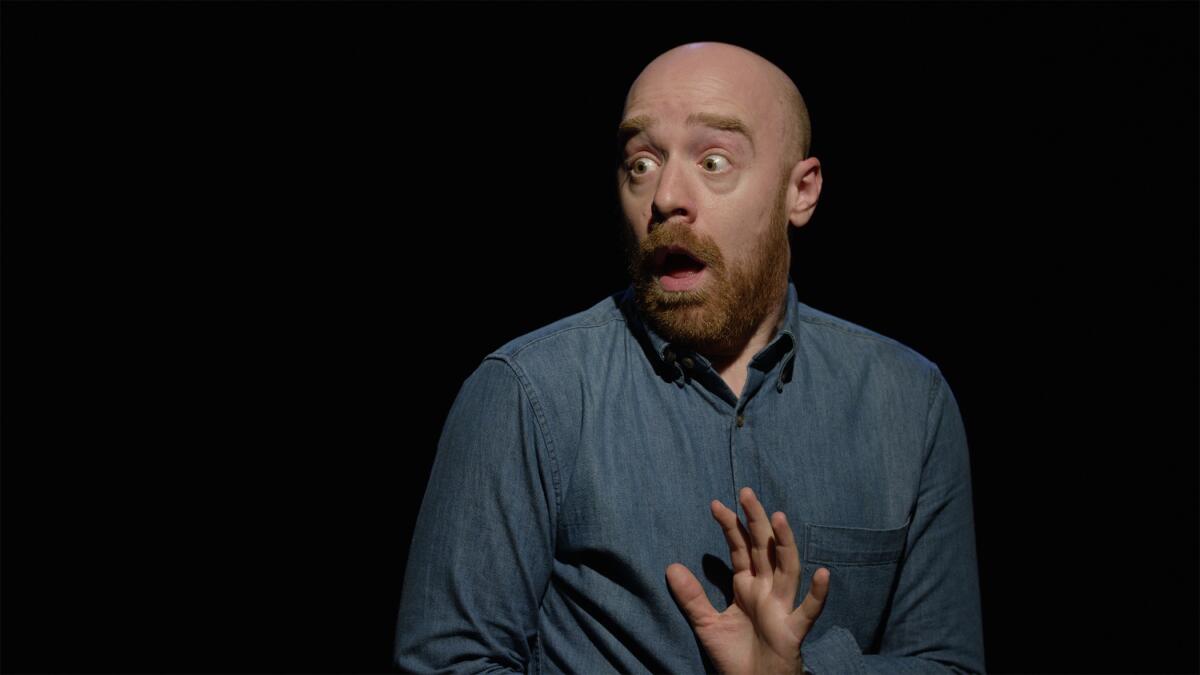 Tom DeTrinis in “Making Friends," holding up his hand while standing in front of a black background. 