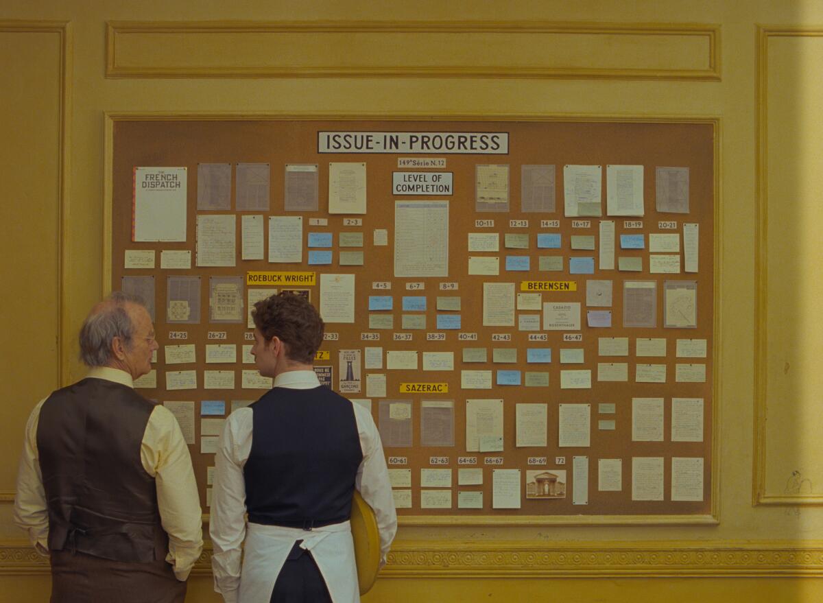 Two actors stand in front of a bulletin board.