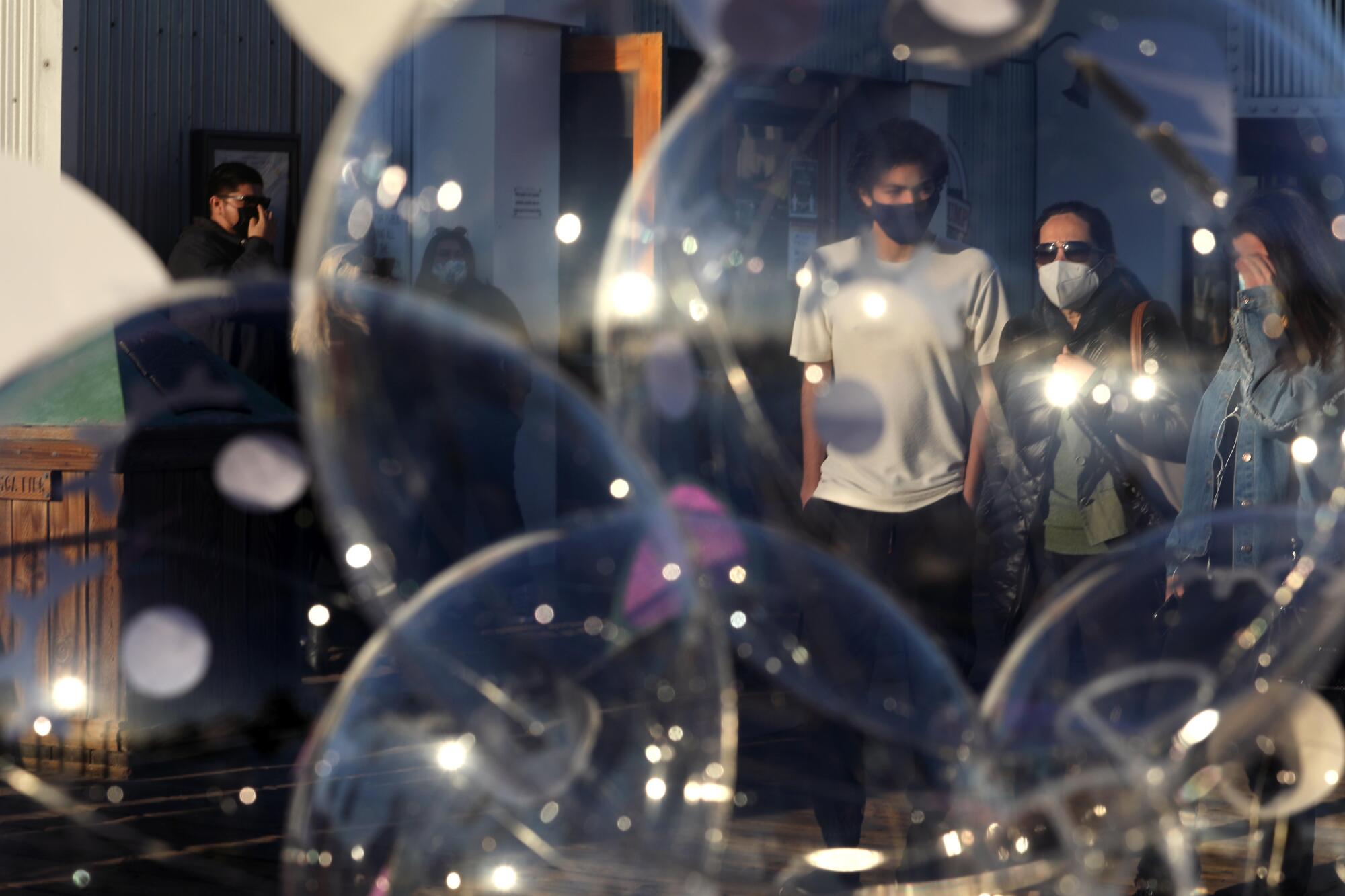 A family wearing masks walks behind a cluster of transparent balloons.