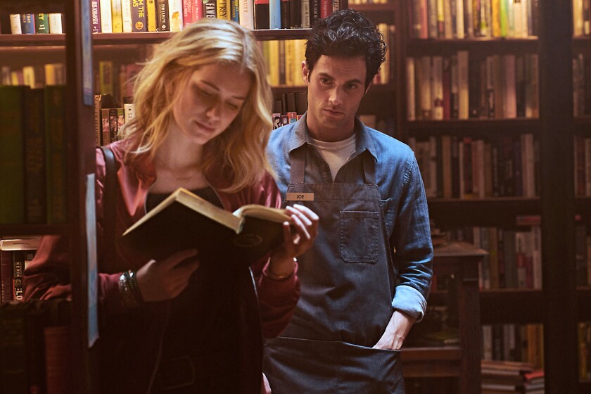 A man watches a woman read a book in front of bookshelves