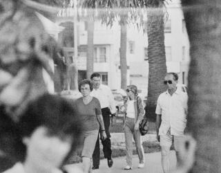 A black-and-white photo of a man in white shirt and dark pants, walking next to a woman, with other people nearby
