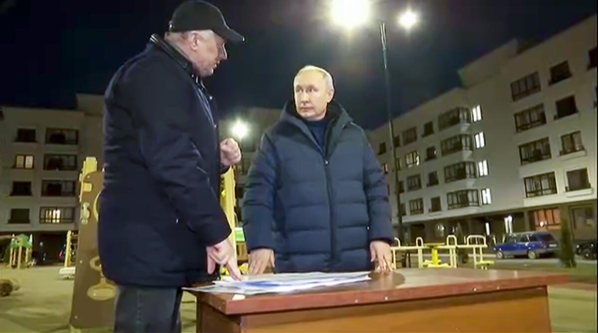 Russian President Vladimir Putin listens to a second man near what looks like a newly built residential complex 