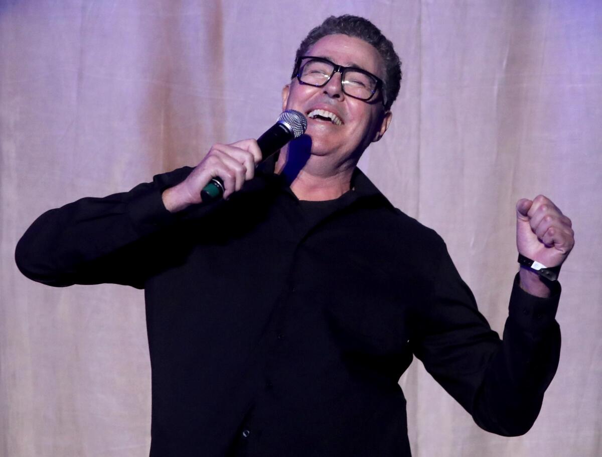 Adam Carolla onstage with his eyes closed, one hand clenched as the other holds a microphone