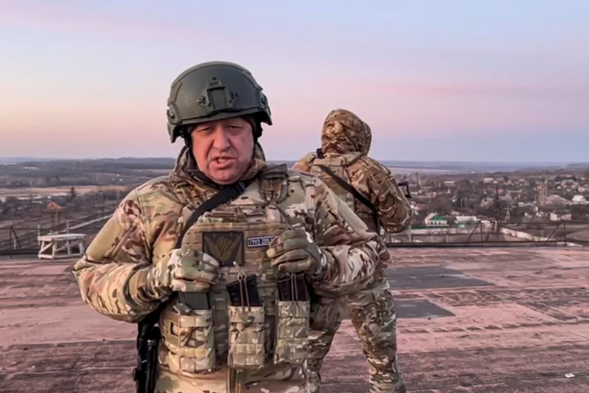 Мужчина лет 60 в каске и военной форме разговаривает. Позади него стоит еще один мужчина в военной форме, смотрящий в другую сторону.