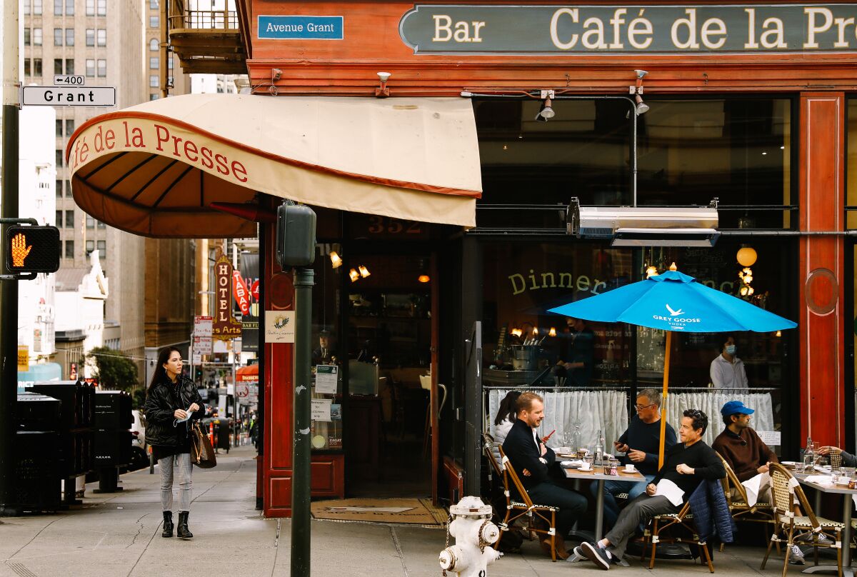 People in jackets sit outside a corner cafe with a curving canopy in a city setting. 
