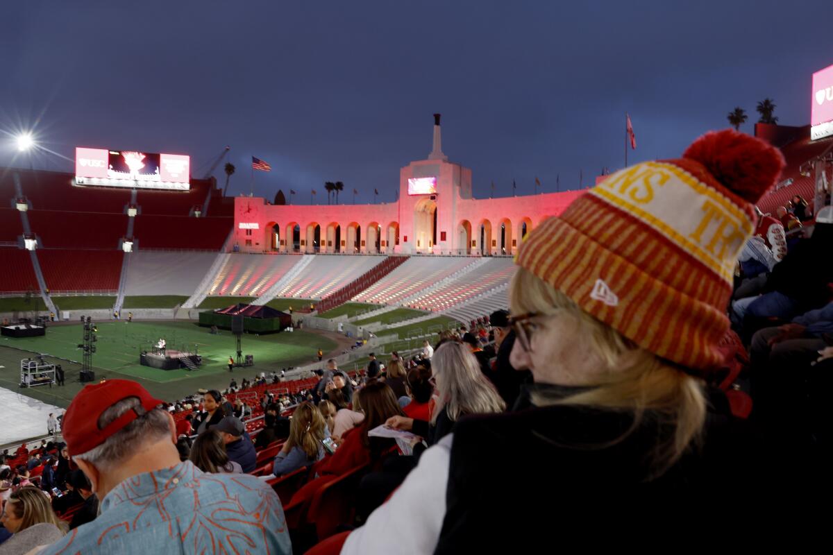 A woman wearing a Trojans cap and sock stands among a small crowd at a large football stadium.