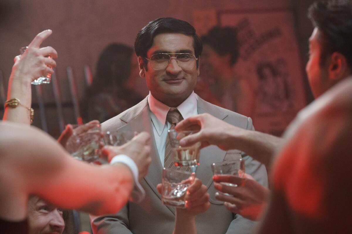 A man in a brown suit and glasses smiles as people toast him.