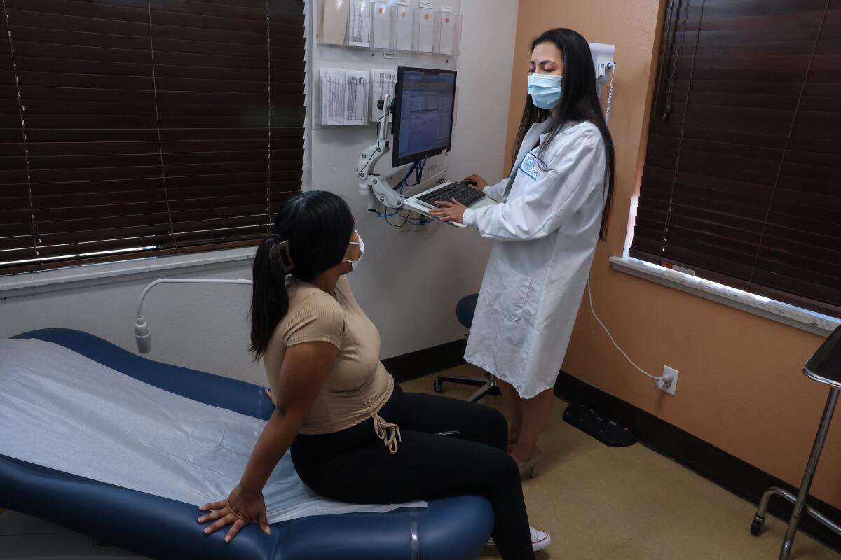 A masked woman in a white coat standing at a workstation speaks to a woman sitting in an examination chair 