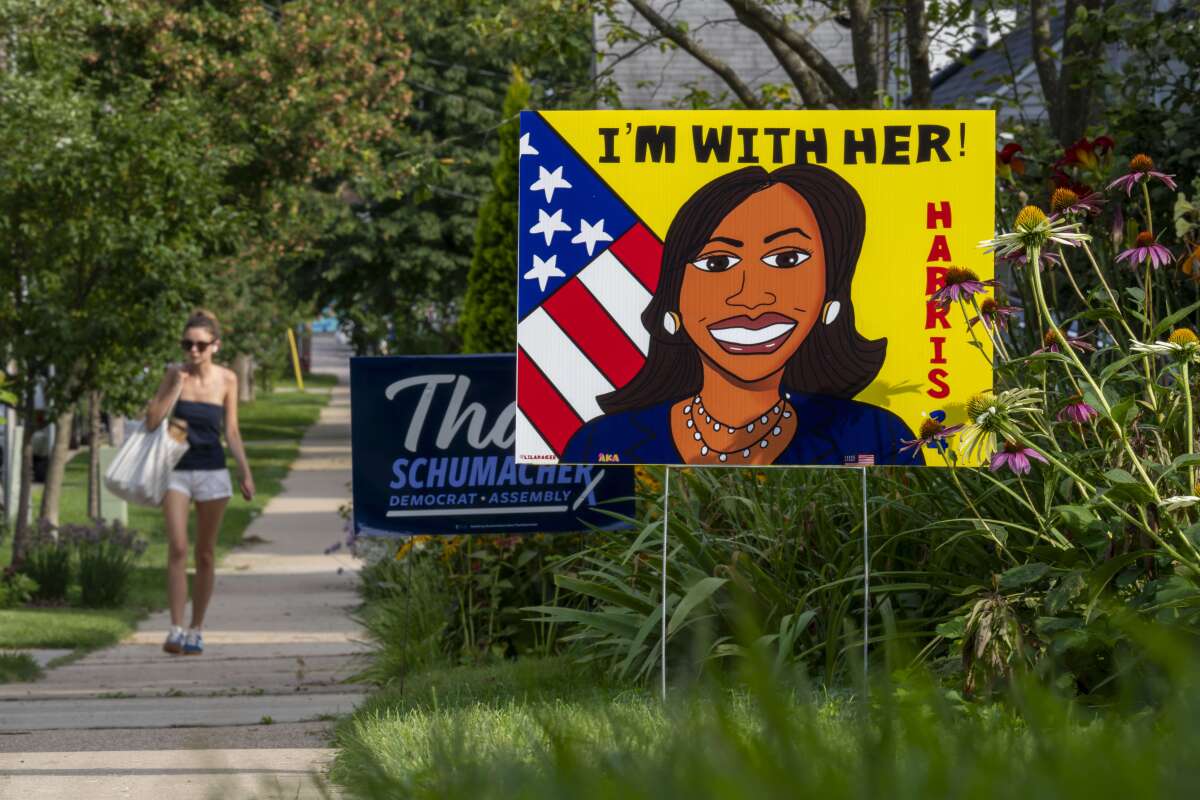 A colorfully painted sign portrays Kamala Harris under the words, "I'm with her," as a pedestrian walks on a passing sidewalk