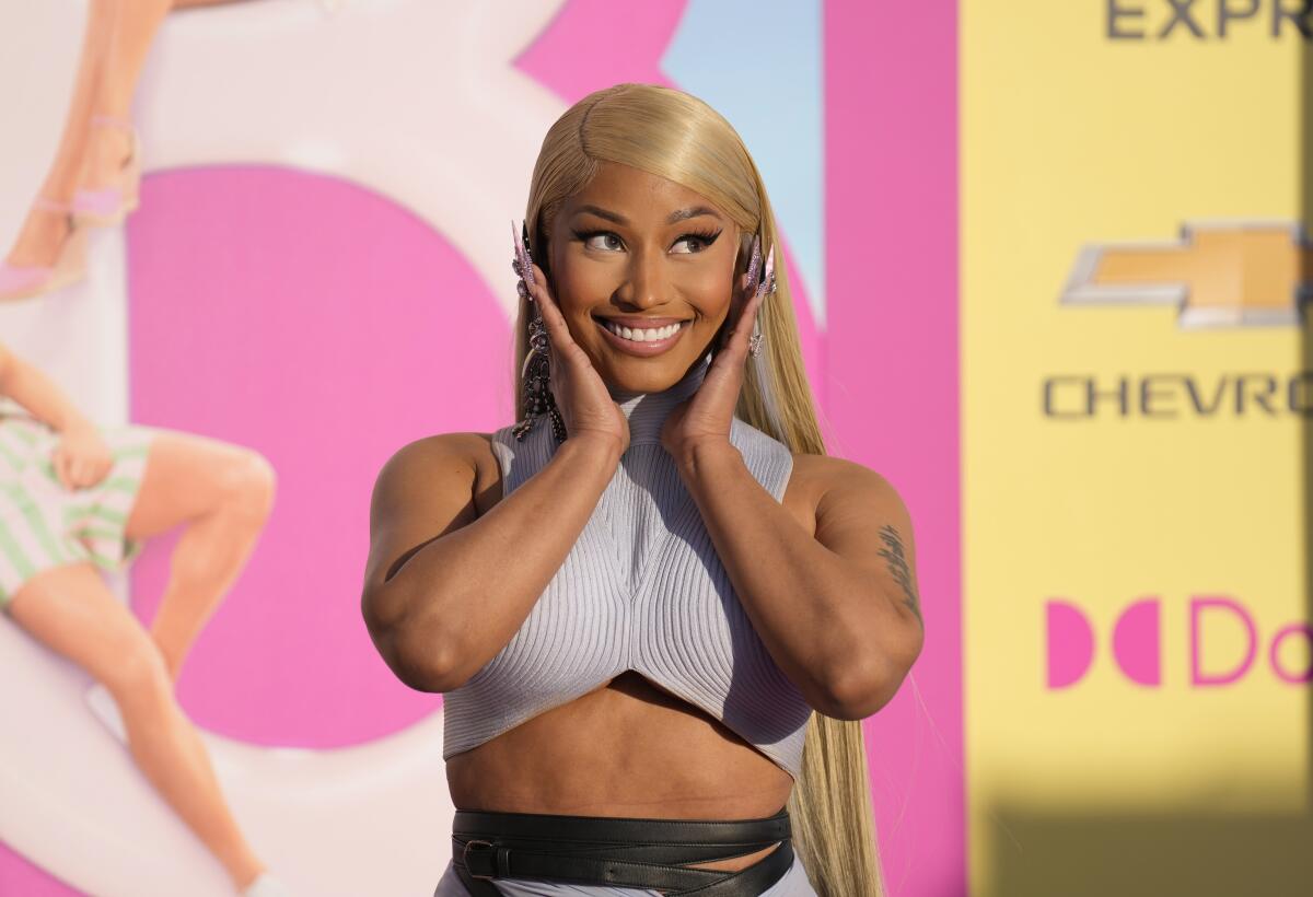 Nicki Minaj in a matching blue-gray top and skirt, holding her hands to her face and smiling in front of a Barbie poster