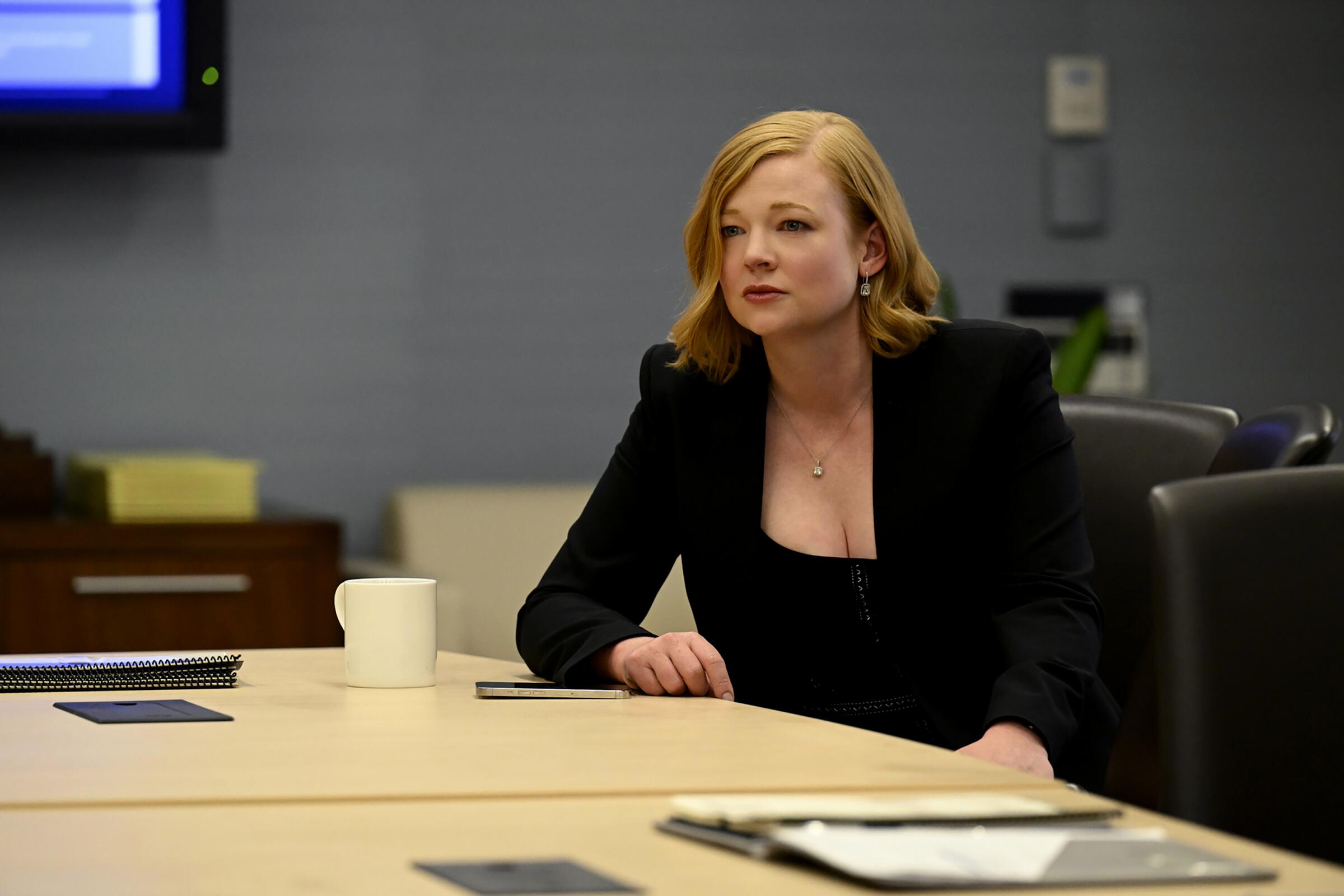 A woman sits at an office table with her phone in front of her in "Succession."
