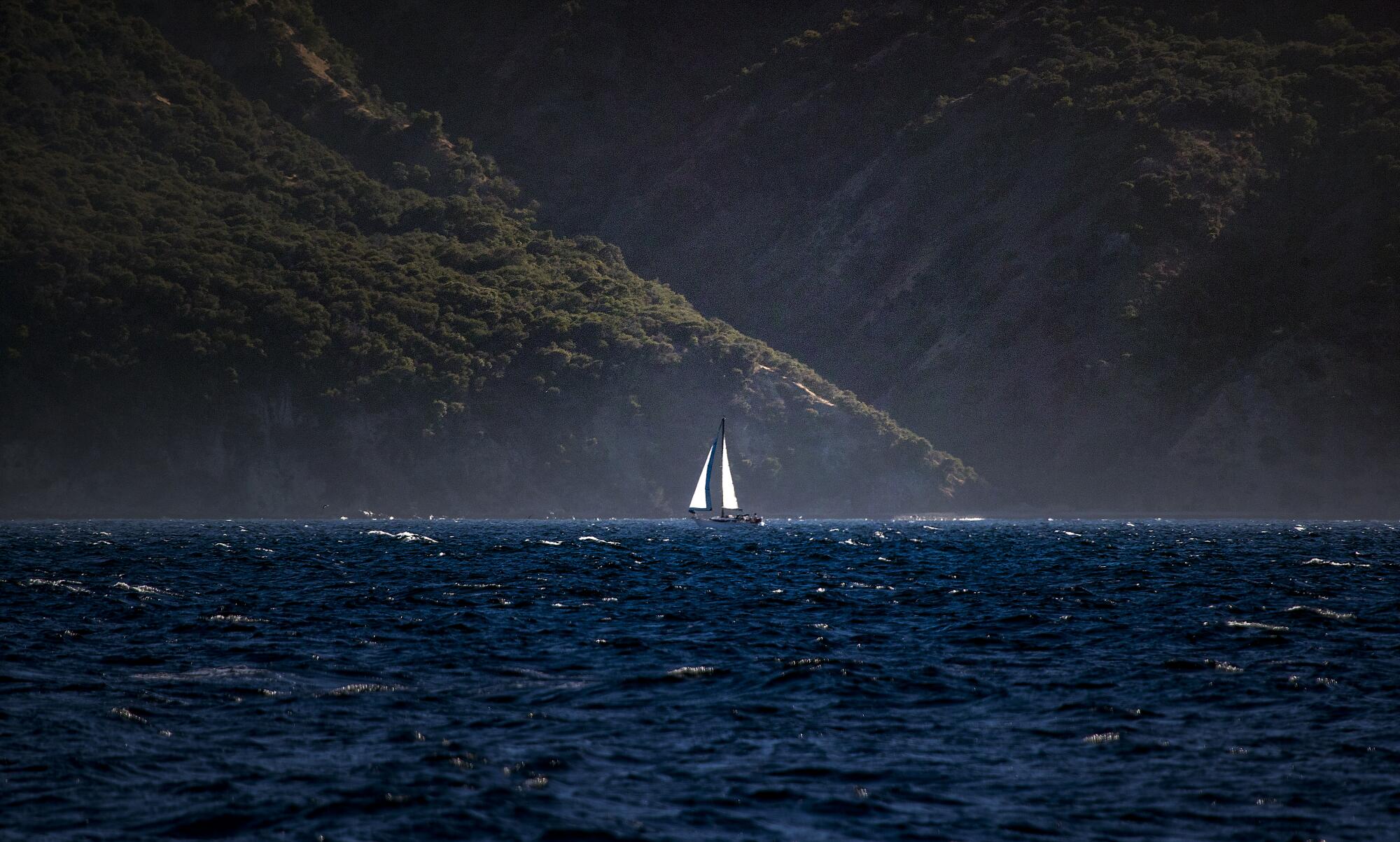 Mountains on an island rise behind a lone sailboat.