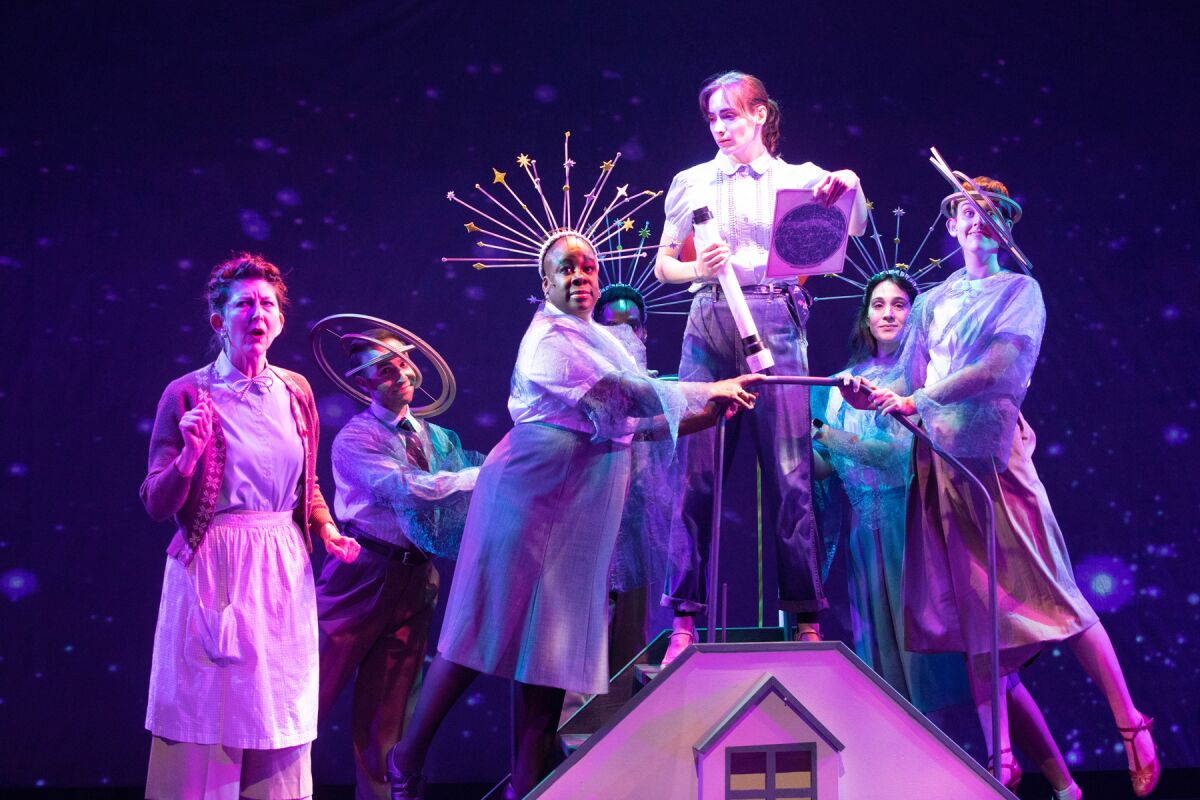 Actors in costume cluster around a woman standing on a model house.