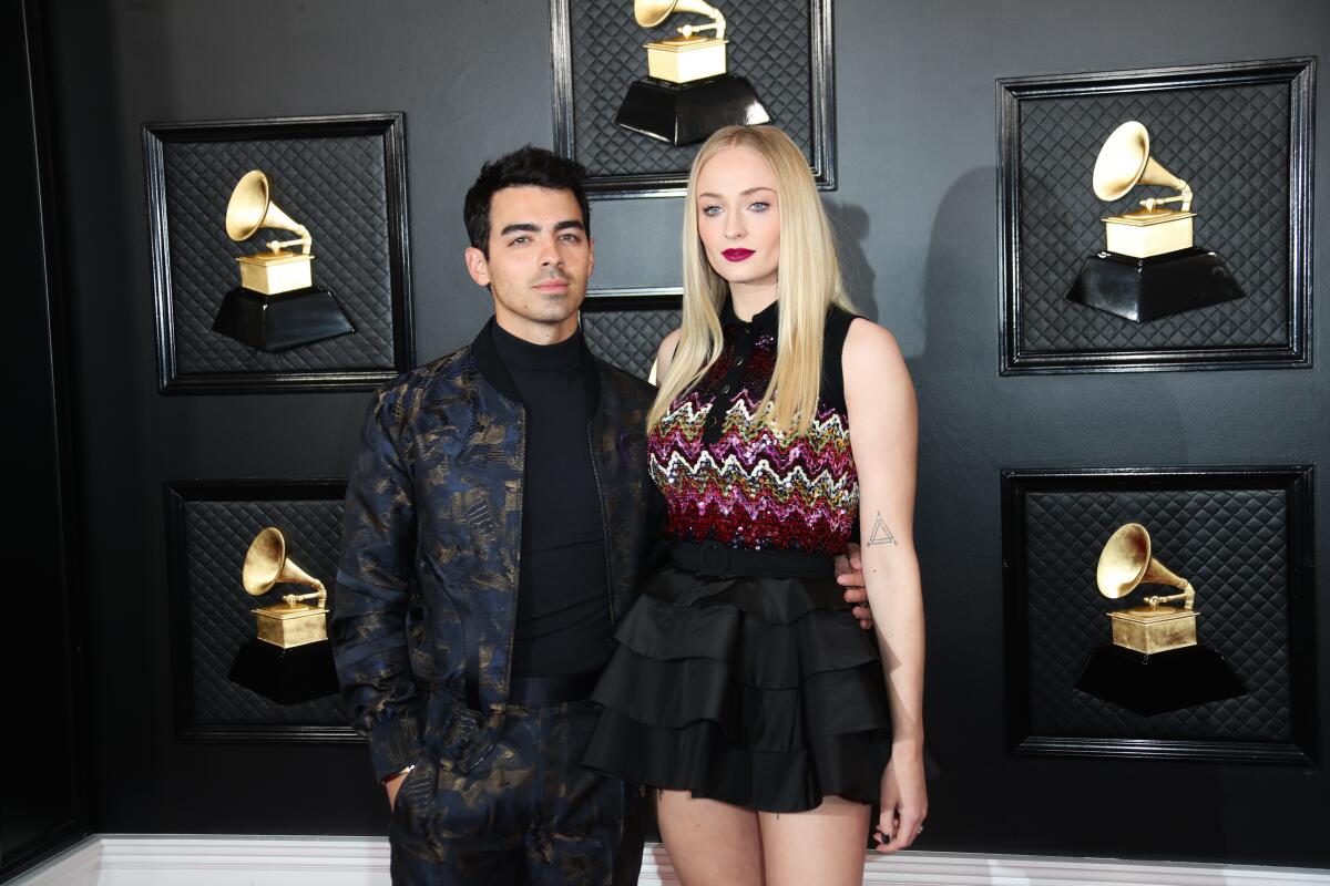 Joe Jonas and Sophie Turner pose together in front of a wall with photos of Grammy awards