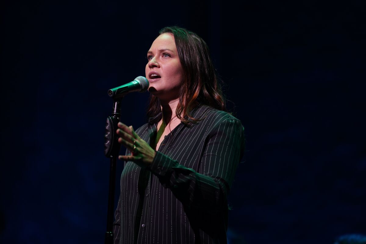A woman in a dark shirt stands at a microphone.