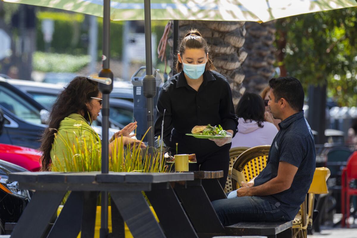 A masked server delivers food to diners at an outdoor restaurant
