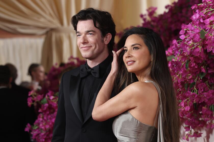 John Mulaney in a black suit posing next to Olivia Munn in a silver gown at the Oscars red carpet